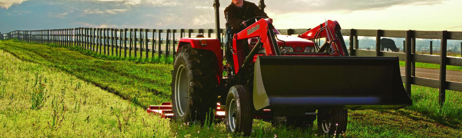 A person riding a 2019 Mahindra 2wd tractor through some grassland next to a fence with a cow in the background.